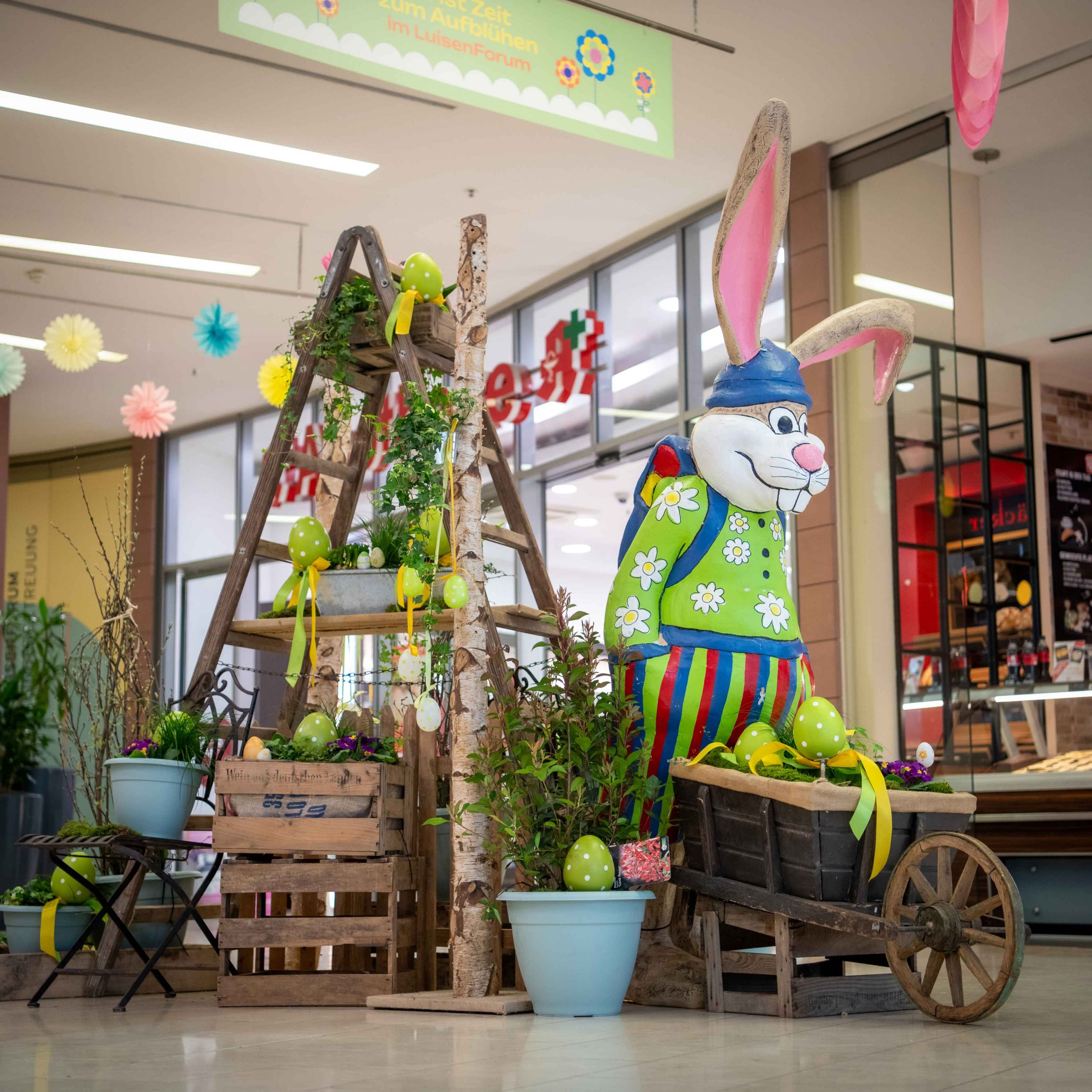 Große bunte Hasenfigur mit Blumenwagen, umgeben von gelber Osterdeko und Pflanzkübeln.