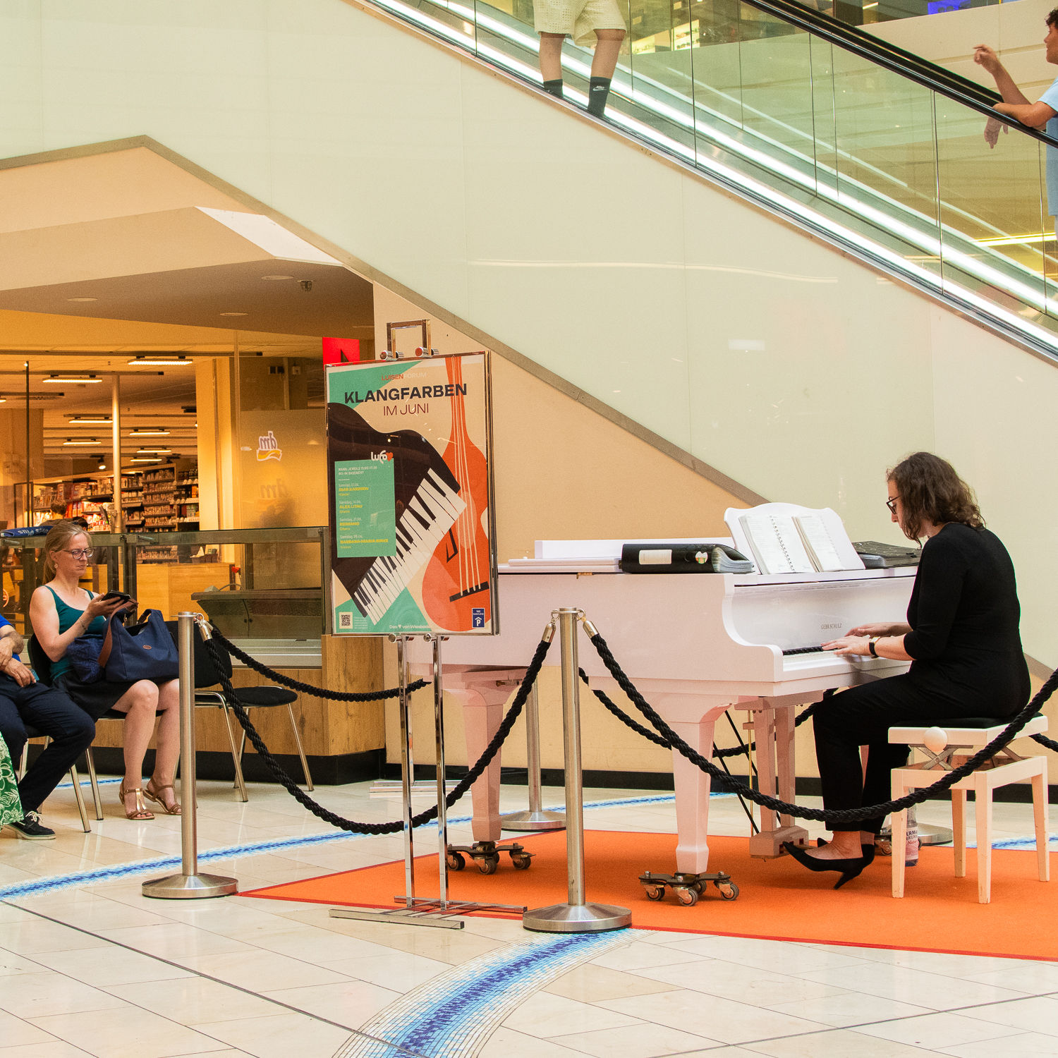 Die Pianistin Barbara-Maria Birke spielt auf einem weißen Flügel im LuisenForum Wiesbaden. Im Hintergrund sind Zuschauer:innen, ein Veranstaltungsplakat und eine Rolltreppe zu sehen.