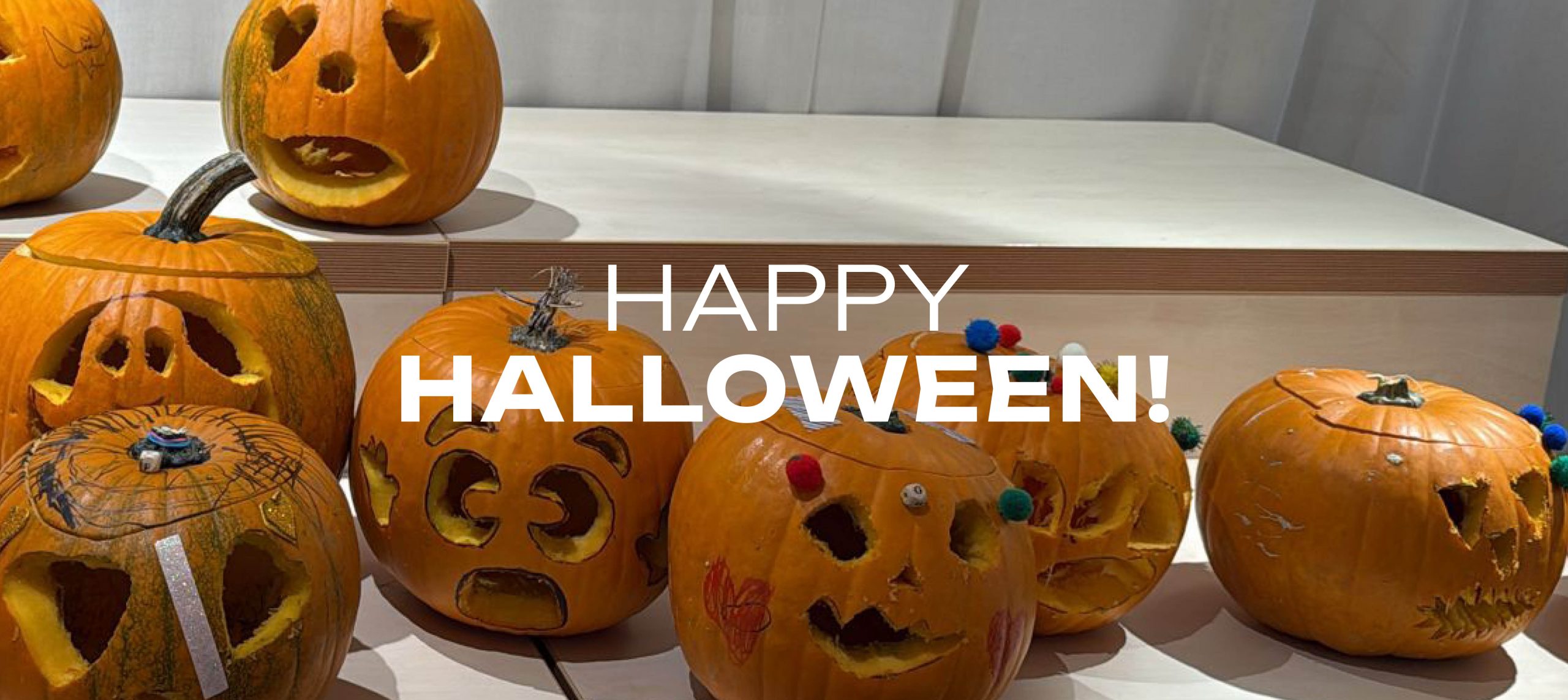 ChatGPT: Mehrere geschnitzte Kürbisse mit verschiedenen Gesichtern stehen auf Tischen vor einem grauen Vorhang, darüber steht der Text „HAPPY HALLOWEEN!“ und daneben ein orangefarbener Stern mit „am 31.10.2025“.